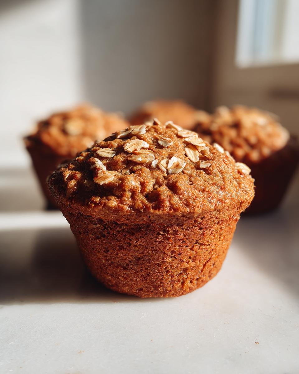 A close-up of a freshly baked banana oatmeal muffin topped with rolled oats, sitting on a white surface.