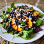 A vibrant blueberry peach feta salad served on a white plate, featuring mixed greens, fresh blueberries, peach slices, feta cheese, pecans, and a balsamic glaze.