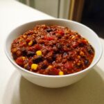 Close-up of a white bowl filled with rich, thick black bean chili featuring visible corn kernels and tomatoes.