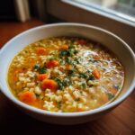 Close-up of a white bowl filled with hot pastina soup, featuring tiny star and tube pasta, sliced carrots, and fresh parsley.