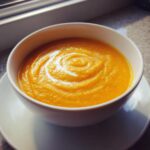 A close-up of a white bowl filled with vibrant orange squash soup, garnished with a swirl of cream.