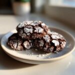 Three rich, dark brown butter toffee chocolate crinkle cookies dusted heavily with powdered sugar, stacked on a small white plate.