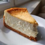 A close-up shot of a perfect slice of creamy cheesecake recipe on a white plate, showing golden top and graham cracker crust.