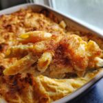 A spoonful of creamy penne mac and cheese lifted from a baked casserole dish, topped with crispy breadcrumbs.