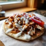 Close-up of open-faced chicken shawarma on pita bread topped with red onion, tomato, and generous garlic sauce.