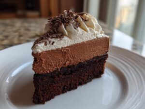 A decadent slice of chocolate mousse cake featuring a dark brownie base, thick chocolate mousse layer, and whipped cream topping with shavings.