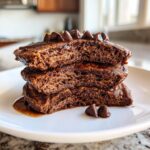 Close-up of a stack of three fluffy chocolate oat pancakes drizzled with syrup and topped with chocolate chips.