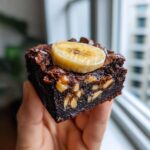 Close-up of a rich, fudgy chunky monkey brownie held in hand, topped with a banana slice and walnuts.