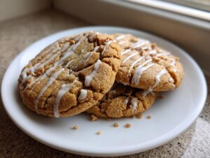 Three soft, crinkled cinnamon toast crunch cookies drizzled with white icing, stacked on a white plate.