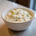 Close-up of a creamy, chunky cheese dip with visible green herbs and peppers served in a small white bowl.