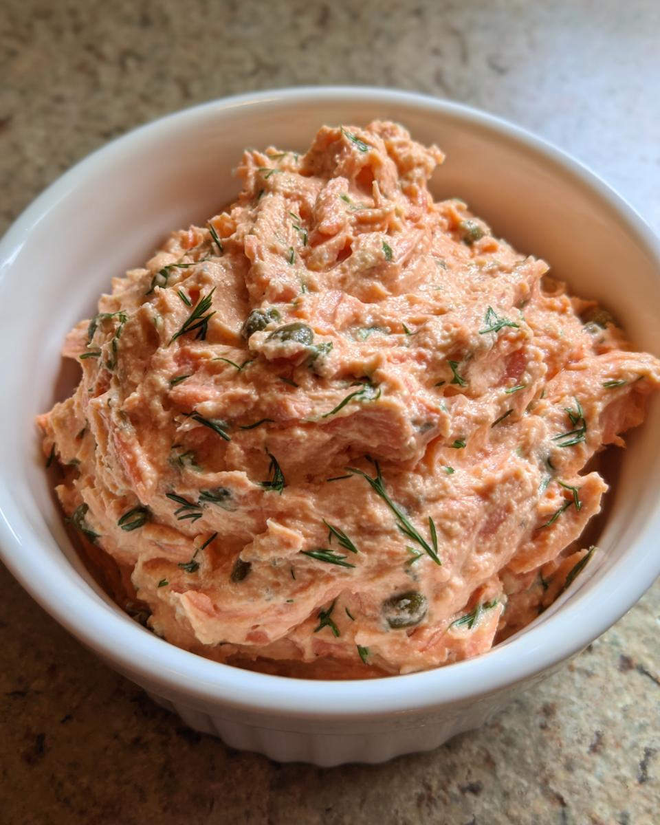 A close-up of creamy, pink smoked salmon dip garnished with fresh dill and capers in a white serving bowl.
