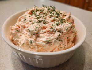 A white bowl filled with creamy, pink smoked salmon dip, generously garnished with fresh dill.
