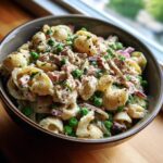 Close-up of a creamy tuna pasta salad mixed with bright green peas and red onion in a dark bowl.