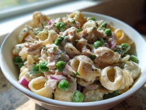 Close-up of creamy tuna pasta salad featuring shell pasta, bright green peas, red onion, and tuna in a white bowl.