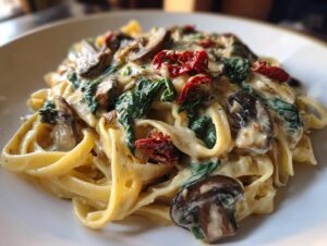 A close-up of creamy Tuscan mushroom pasta with spinach and sun-dried tomatoes coating fettuccine noodles.