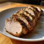 Slices of perfectly cooked crockpot pork tenderloin, seasoned heavily with herbs and resting in its juices on a white plate.