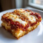 Close-up of a square slice of baked eggplant parmesan with crispy breading, rich tomato sauce, and melted mozzarella cheese.