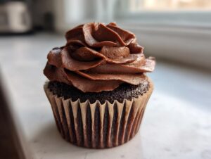 A single, rich chocolate cupcake topped with a generous swirl of dark chocolate frosting, sitting on a white counter.