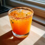 Close-up of a bright orange firecracker margarita in a rocks glass with a chili salt rim and visible jalapeño pieces.