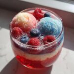 A colorful glass of firecracker punch showing red, white, and blue layers, topped with scoops of sherbet and fresh raspberries and blueberries.