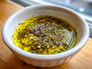 Close-up of a white bowl filled with vibrant olive oil bread dip, featuring green herbs and red pepper flakes.