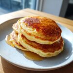 A stack of three perfectly golden greek yogurt pancakes drizzled generously with maple syrup on a light blue plate.
