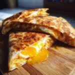 Two halves of a golden brown fried egg quesadilla cut open, showing a runny yolk spilling onto a wooden board.