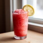 A tall glass filled with bright pink, icy frozen strawberry lemonade, garnished with a lemon slice.