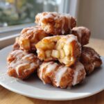 A stack of golden brown apple fritter bites drizzled with white icing, one is broken open showing diced apple filling.
