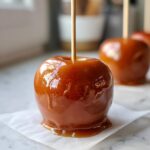 Close-up of a perfectly coated, glossy caramel apple on white parchment paper, ready to eat.