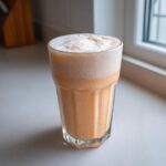 A tall, faceted glass filled with a thick, orange-hued high-protein peaches n cream protein milkshake topped with a layer of white foam.