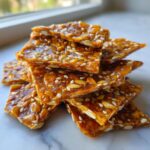 A stack of glossy, amber-colored pieces of homemade seed brittle featuring sunflower seeds and sesame seeds.