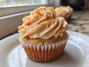 A close-up of a single honey peach cream cheese cupcake with swirled frosting and crumbs on a white plate.