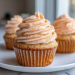 A close-up of one perfectly frosted honey peach cream cheese cupcake sitting on a white plate.