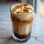 Close-up of a tall glass containing an iced caramelized banana latte with thick, swirled caramel foam on top.