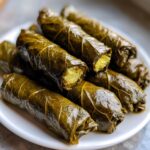 Close-up of several perfectly rolled lemony rice-stuffed grape leaves stacked on a white plate.