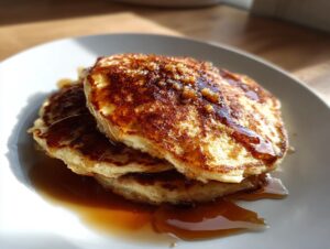 A stack of three golden brown maple ginger pancakes generously drizzled with syrup on a white plate.