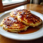 Three golden brown maple ginger pancakes stacked on a white plate, generously drizzled with syrup and candied ginger pieces.