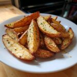 A close-up of crispy, golden mediterranean potato wedges seasoned with herbs served on a white plate.