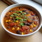 Close-up of a white bowl filled with rich, red Mexican quinoa soup, featuring black beans, corn, and fresh cilantro.