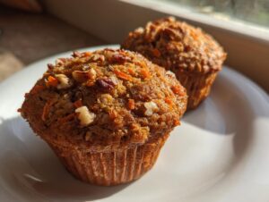 Close-up of two freshly baked morning glory muffins topped with shredded carrots and chopped pecans on a white plate.