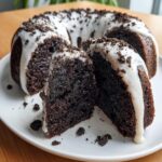 Close-up of a rich, dark chocolate Oreo Bundt Cake with white vanilla glaze and cookie crumbs, two slices cut out.