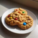 Three freshly baked peanut butter m&m cookies stacked slightly on a white plate, illuminated by sunlight.