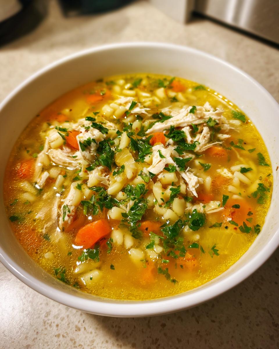 Close-up of a warm bowl of penicillin soup featuring shredded chicken, small pasta, carrots, and fresh parsley garnish.