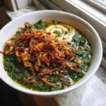 A close-up of a bowl of rich, green Persian noodle soup topped with crispy fried onions and a swirl of yogurt.