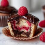 Close-up of a raspberry chocolate cupcakes cut in half, showing marbled cake, raspberry filling, and chocolate ganache topped with a fresh raspberry.