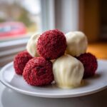 A stack of homemade red velvet cake balls coated in crumbs next to white chocolate-dipped truffles on a white plate.