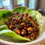 A close-up of a single serving of savory chicken lettuce wraps filling, featuring glossy sauce, chicken pieces, and nuts, nestled in a crisp lettuce cup.
