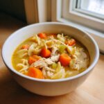 A close-up of a white bowl filled with rich, yellow slow cooker chicken noodle soup featuring shredded chicken, egg noodles, and large chunks of carrots.