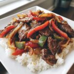 A close-up of tender slow cooker pepper steak mixed with red and green bell peppers and onions, served over a bed of white rice on a square white plate.